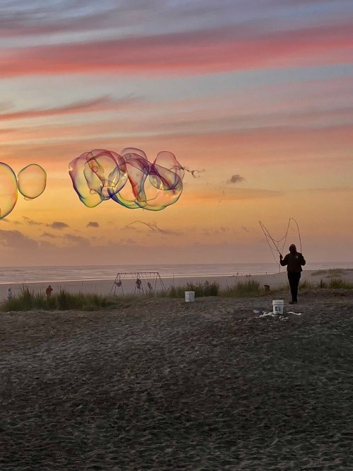A person creates large bubbles against a colorful sunset at the beach, with swings and people in the background.