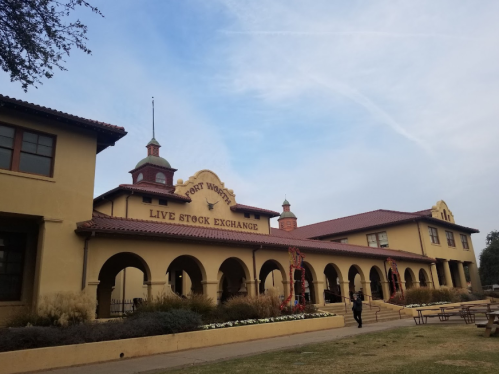 Historic building with arched windows and a sign reading "Fort Worth Live Stock Exchange," surrounded by greenery.