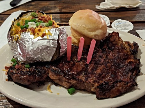 A plate featuring a grilled steak, a loaded baked potato, and a biscuit, garnished with green onions.