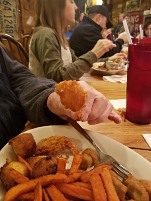 A hand holding a fried food item over a plate of sweet potato fries, with diners in the background.