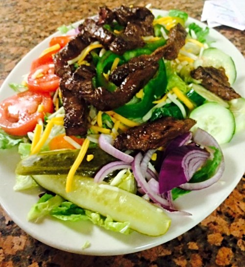 A colorful salad topped with grilled steak, tomatoes, cucumbers, green peppers, red onions, and shredded cheese.
