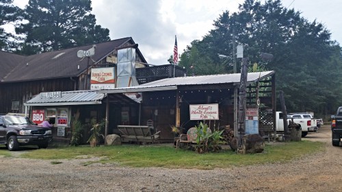A rustic building with a porch, signs for BBQ and a restaurant, surrounded by trees and parked cars.