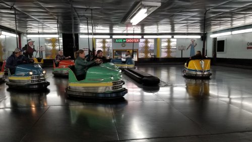 A colorful bumper car arena with people enjoying rides in green, blue, and yellow cars on a shiny floor.