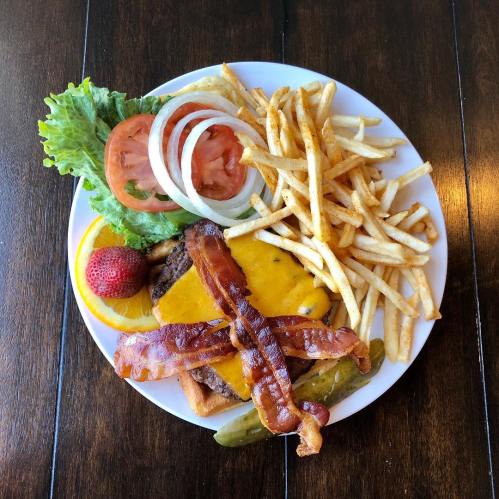 A plate with a cheeseburger topped with bacon, lettuce, tomato, pickles, fries, and a strawberry on the side.