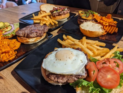 A table with four gourmet burgers, fries, and a side salad, featuring various toppings and a sunny-side-up egg.