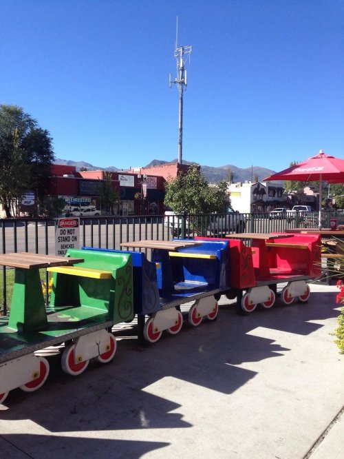 Colorful wooden benches shaped like train cars lined up outside, with a clear blue sky and buildings in the background.
