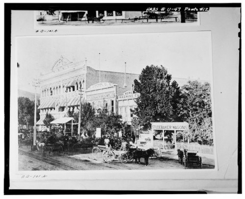 Historic black-and-white photo of a busy street scene with horse-drawn wagons and a large building in the background.