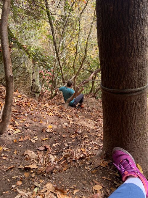 A person sits on a forest floor surrounded by trees and autumn leaves, with a close-up of a foot in the foreground.