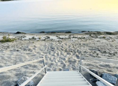 A sandy beach with gentle waves, viewed from white stairs leading down to the water's edge at sunset.