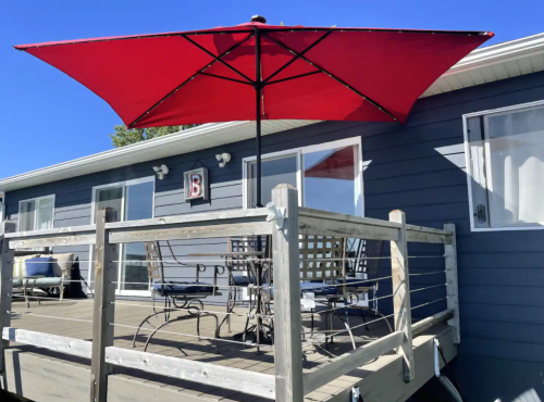 A red umbrella shades a patio with metal furniture on a wooden deck of a blue house under a clear sky.