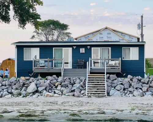 A blue cottage with a porch, surrounded by rocks, near a sandy beach and trees in the background.