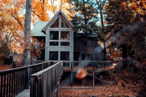 A cozy cabin surrounded by autumn trees, featuring large windows and a wooden walkway leading to the entrance.