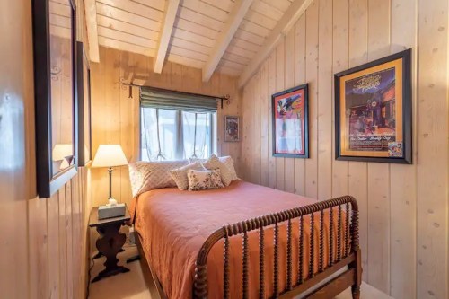 Cozy bedroom with wooden walls, a bed with an orange cover, a lamp, and framed artwork on the walls.
