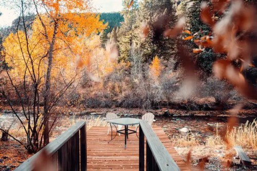 A wooden deck by a river surrounded by vibrant autumn foliage and a table with chairs.
