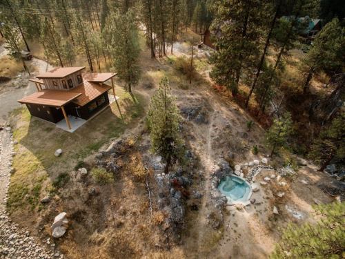 Aerial view of a modern cabin surrounded by trees, with a small hot tub nearby and a rocky landscape.