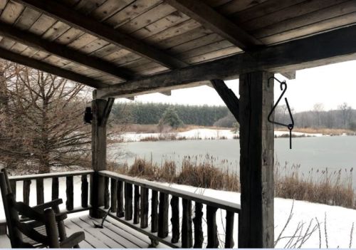 A snowy porch view overlooking a frozen lake and wooded area, with rustic wooden beams and a rocking chair.