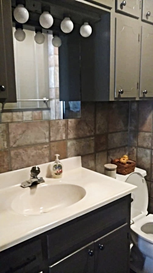 A bathroom with a sink, mirror, and toilet, featuring gray cabinets and tiled walls. A basket of fruit is visible.