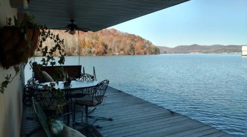 A serene lakeside view from a porch, featuring a table and chairs, with autumn foliage on the opposite shore.
