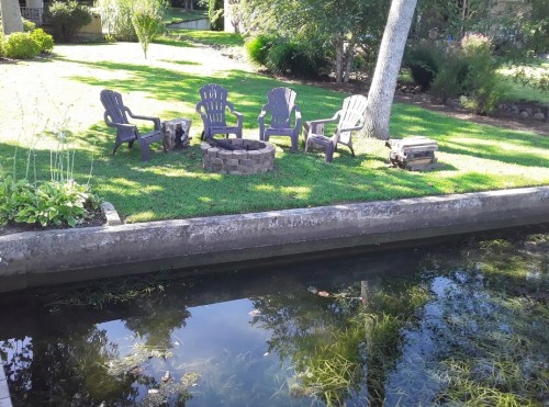 A serene backyard scene featuring four purple chairs around a stone fire pit by a calm waterway.