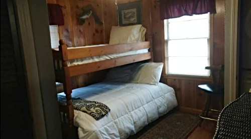 Cozy bedroom with a wooden bunk bed, a double bed, and a window, featuring rustic wood paneling and warm decor.