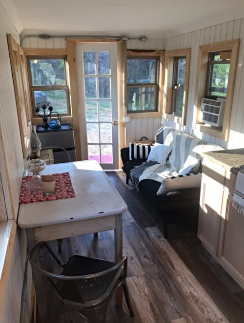 Cozy interior of a small cabin with a table, chairs, and a sofa, featuring large windows and natural light.