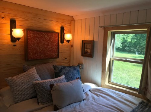 Cozy bedroom with a wooden wall, decorative pillows on a bed, and a window overlooking a green landscape.