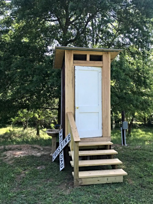 A wooden outhouse with a white door, set on steps, surrounded by greenery and a "CROSSING" sign nearby.