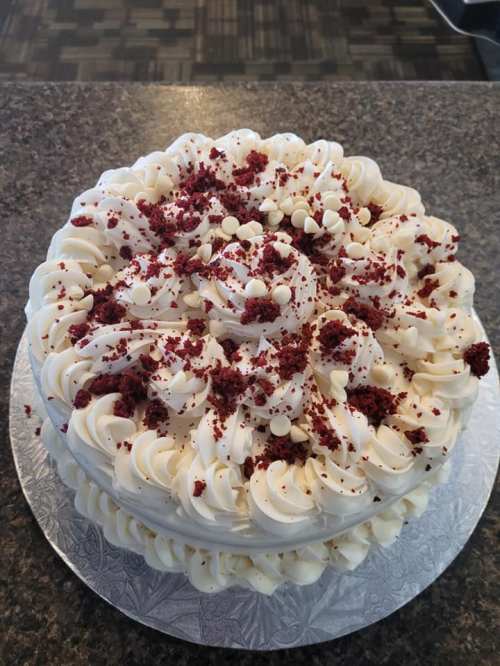 A round cake with white frosting, red velvet crumbs, and white chocolate chips on top, displayed on a silver cake board.