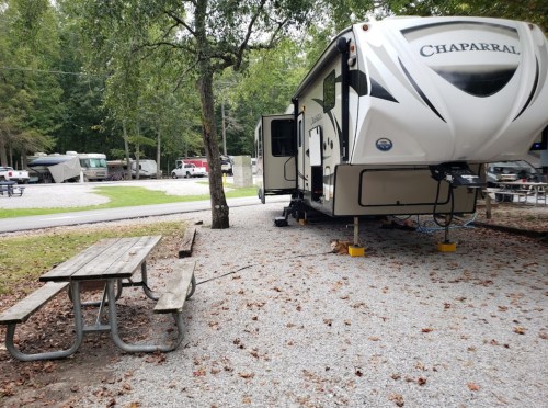 A parked RV in a campground with a picnic table nearby, surrounded by trees and gravel.
