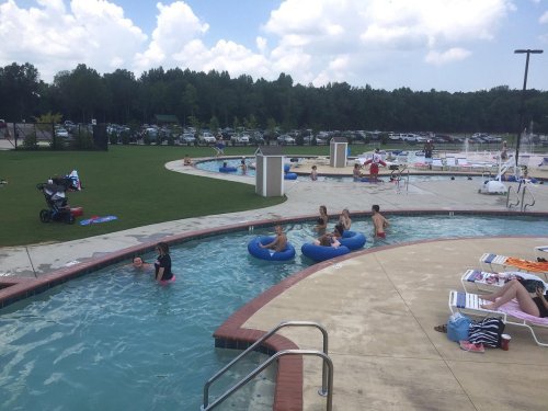 A pool area with people relaxing on blue floats, sunbathing, and enjoying the water under a partly cloudy sky.