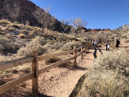 A dirt path lined with a wooden fence, surrounded by sparse vegetation and rocky hills, with people walking in the distance.