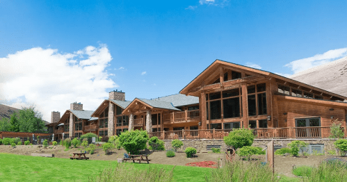 A large wooden lodge with multiple gabled roofs, surrounded by green grass and shrubs under a blue sky.