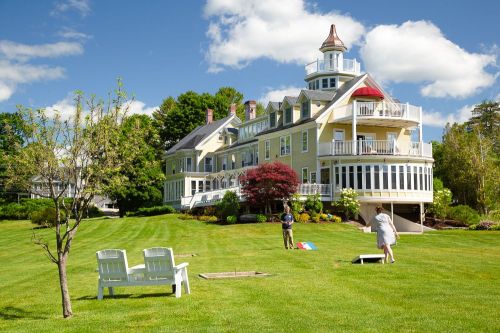 A large yellow house with a tower, surrounded by green lawn and trees, with people playing games in the foreground.