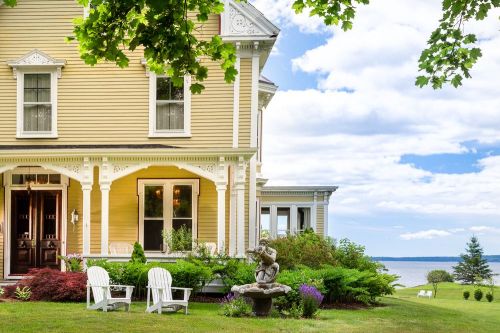 A charming yellow house with a porch, surrounded by greenery and a view of the water under a partly cloudy sky.