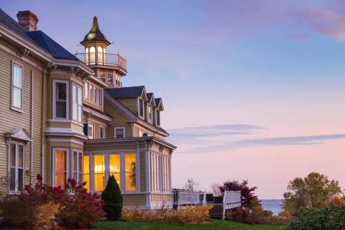 Victorian-style house with a lighthouse tower, surrounded by colorful foliage at sunset.