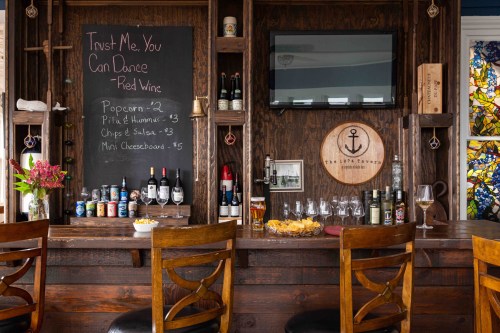 A cozy bar with a chalkboard menu, wine bottles, and snacks on the counter, featuring wooden stools and a TV.