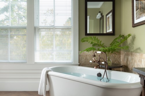 A bright bathroom featuring a freestanding tub, a fern, and large windows with blinds, creating a serene atmosphere.