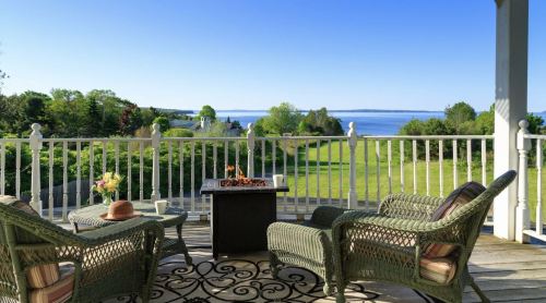 A serene porch with wicker chairs, a fire pit, and a view of a calm lake and green landscape under a clear blue sky.