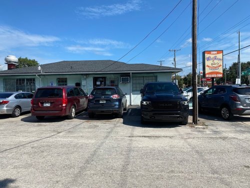 A small building with a sign, surrounded by parked cars on a sunny day.
