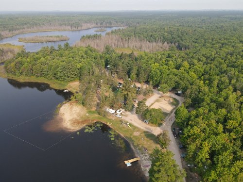 Aerial view of a forested area with a lake, sandy shore, and scattered campsites along the water's edge.