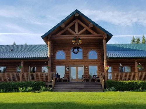 A large log cabin with a green metal roof, featuring a front porch and decorative wreaths, surrounded by greenery.