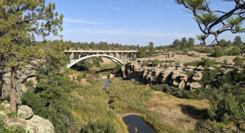 A scenic view of a bridge arching over a rocky landscape with greenery and a winding stream below.