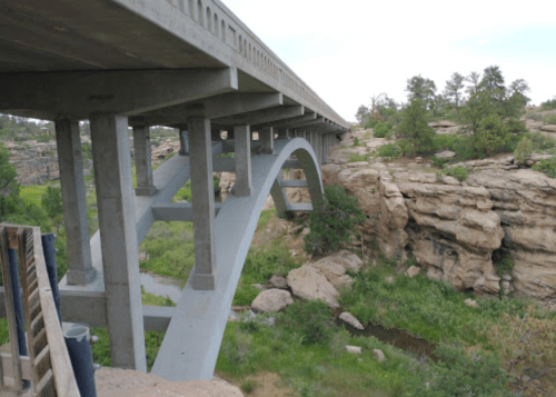 A concrete arch bridge spans over a rocky landscape with greenery below, surrounded by trees and a cloudy sky.