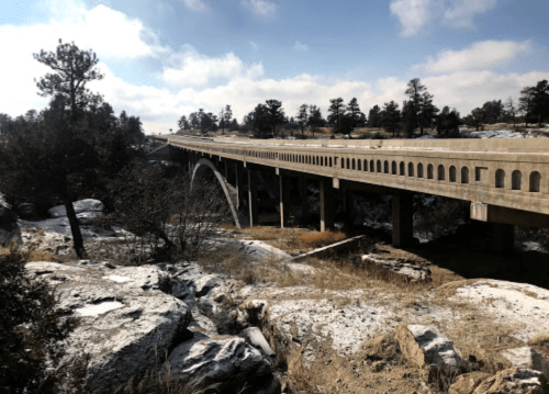 A concrete bridge arches over rocky terrain, surrounded by trees and a cloudy sky.