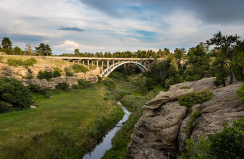 A scenic view of a bridge arching over a lush green valley with a stream and rocky terrain.