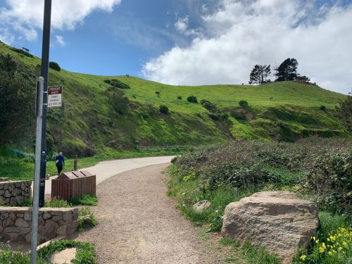 A winding path leads through lush green hills under a partly cloudy sky, with a few people in the distance.
