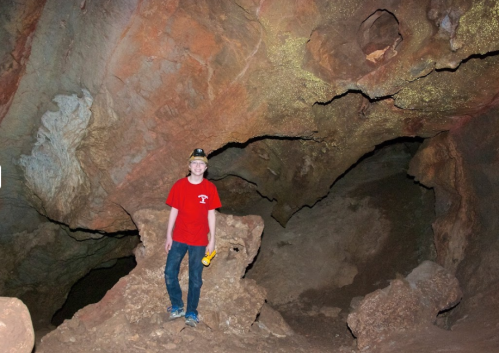 A person in a red shirt stands on rocks inside a cave, holding a flashlight, with textured walls in the background.