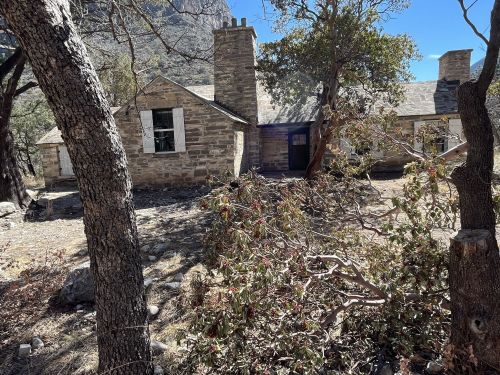 A stone house with a chimney, surrounded by trees and dry vegetation, set against a rocky landscape.