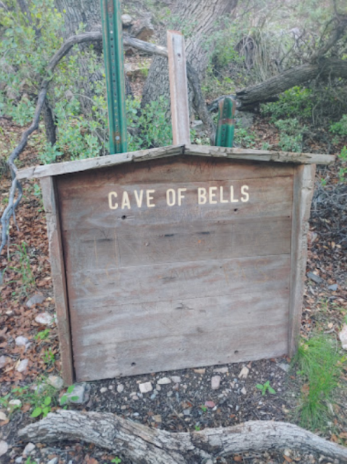 Wooden sign reading "Cave of Bells" in a natural outdoor setting with trees and scattered rocks.