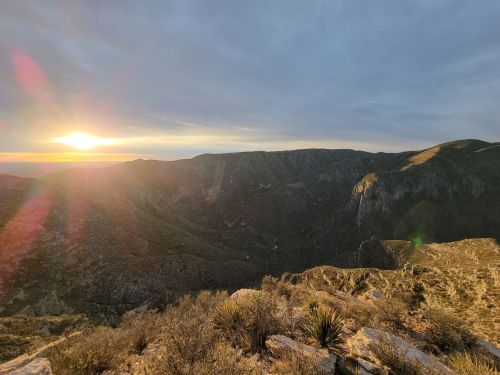 Sunset over a mountainous landscape, with rocky terrain and sparse vegetation under a cloudy sky.